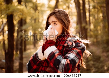 Young woman with nose wiper near autumn tree. Sick girl with runny nose and fever. Showing sick woman sneezing at autumn park. Young woman having flu and blowing her nose