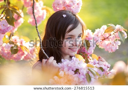 Small brunette smiling girl looking away standing amid pink japanese cherry flowering in broad daylight in the park, horizontal picture