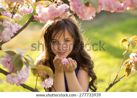 Small brunette girl looking forward smelling cherry bloom standing among pink japanese cherry blossom in broad daylight in the park, horizontal picture