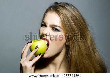 Cruel sexy blonde girl with bright make up looking forward biting fresh green apple standing on gray background copyspace, horizontal picture