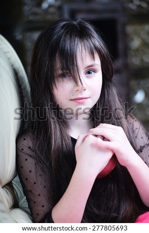 Pretty thoughtful little girl holding red apple sitting on couch looking forward, vertical picture