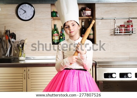 Cute scullion girl in chef\'s hat in the kitchen holding rolling-pin looking forward, horizontal picture
