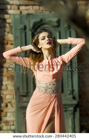 Pretty attractive woman standing near brick wall building looking forward in long peach evening dress outdoor, vertical picture