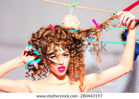 Crazy playful young beautiful fashionable woman with curly hair holding scissors cutting curle looking forward indoor on grey wall background copyspace, horizontal picture