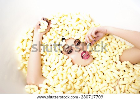 Portrait of sensual young beautiful girl face in corn sticks heap in bathtub with raised hands tasting and  looking forward lying on light food background copy space, horizontal picture