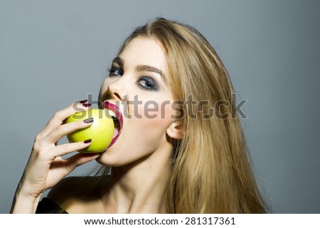 Sexual blonde girl with bright make up looking forward biting fresh green apple standing on gray background copyspace, horizontal picture