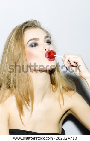 Sexy blonde girl portrait with closed eyes with bright make up holding and licking round red sugar candy standing on light gray background copyspace, vertical picture