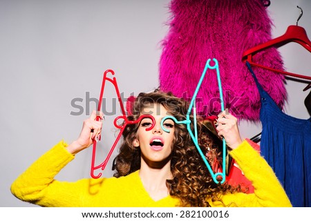 Crazy young girl with curly hair in yellow sweater holding hangers standing amid colorful clothes pink red blue colors on grey wall background, horizontal picture