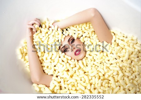 Portrait of pretty young beautiful girl face in corn sticks heap in bathtub with raised hands looking forward lying on light food background copy space, horizontal picture