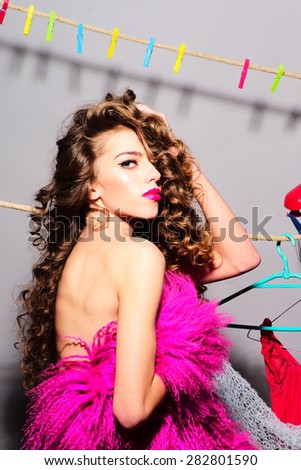 Pretty young sophisticated girl with bright make up holding pink fur cloth looking forward touching her curly hair standing on grey wall background, vertical picture