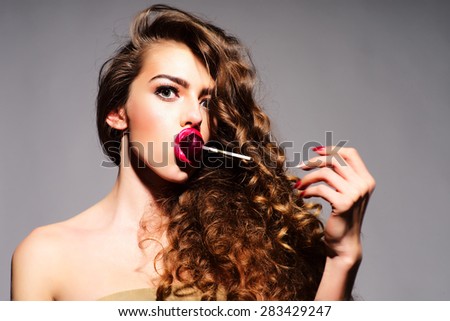 Taking playful young woman with curly hair and bright pink lips holding purple round lollipop in mouth looking forward with big eyes standing on grey background copy space, horizontal picture