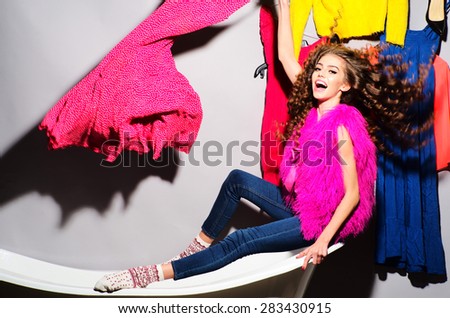 Crazy emotional young woman with curly hair in pink fur vest and blue jeans sitting in white bathtub amid colorful clothes pink orange red blue colors on grey wall background, horizontal picture