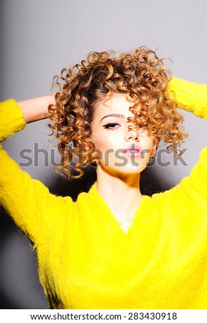 Portrait of pretty young girl touching her curly hair in yellow sweater looking forward standing on grey wall background, vertical picture