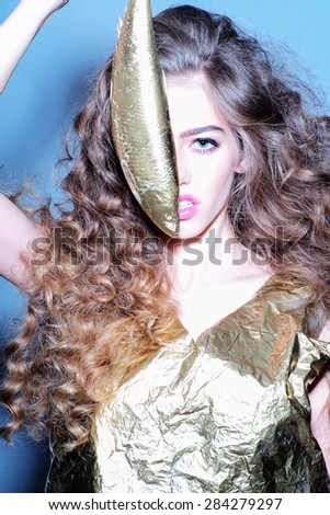 Alluring young girl with curly hair in gold jacket holding golden fish covering half face looking forward standing on grey blue  background, vertical picture
