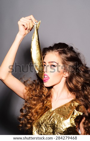 Cute young girl with curly hair in gold jacket holding and licking golden fish looking forward standing on grey background, vertical picture