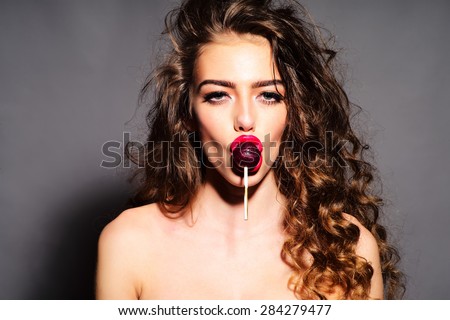 Delightsome young undressed woman with curly hair and bright pink lips holding purple round lollipop in mouth looking forward standing on grey background, horizontal picture