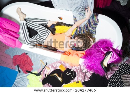 Showy cool young woman with curly hair lying in white bathtub amid colorful clothes pink orange red blue colors on grey wall background, horizontal picture