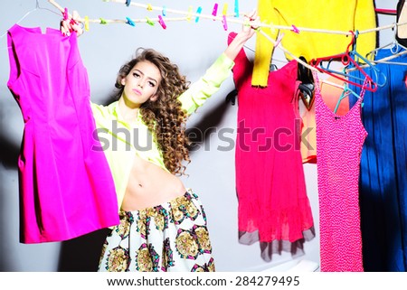 Sensual pretty young girl with curly hair in skirt and blouse standing amid colorful clothes pink orange red blue colors on grey wall background, horizontal picture