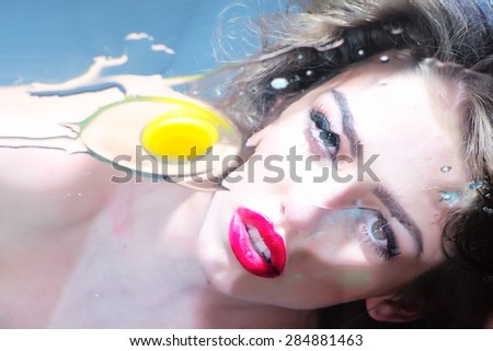Adorable pretty cute girl with curly hair big beautiful eyes and bright pink lips looking through the flat glass surface with raw yellow yolk and white of an egg on blue background, horizontal picture