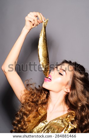 Pretty young girl with curly hair in gold jacket holding and licking golden fish standing on grey background, vertical picture