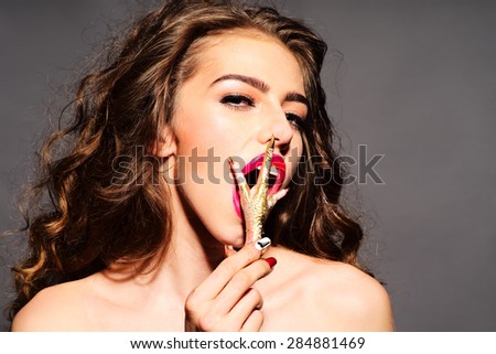 Captivating young undressed woman with curly hair and bright pink lips holding gold chicken foot near open mouth looking forward standing on grey background copy space, horizontal picture