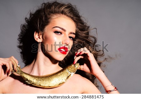 Sensual young naked girl with curly hair holding golden fish as necklace looking forward standing on grey background, horizontal picture