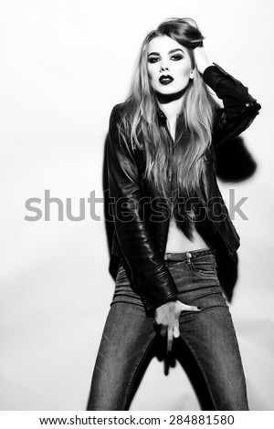 Tough seductive young blonde woman black and white colors with bright make up in leather jacket and jeans looking forward stanging on light background copyspace, vertical picture