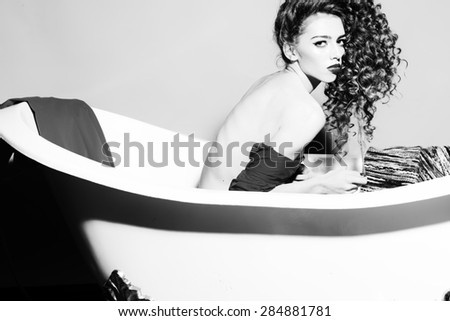 Naked sensual bright young woman with curly hair looking forward sitting in bath on wall background black and white, horizontal photo