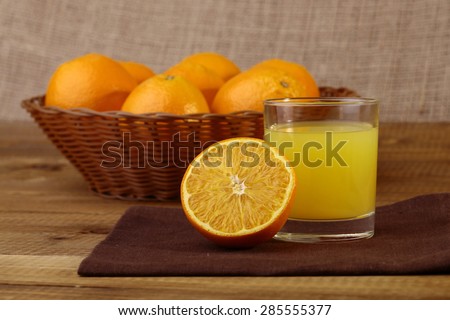 Half orange fruit and glass of yellow juice on wooden table top with basket full of oranges in the background, horizontal picture