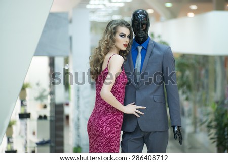 Sexy young girl with bright makeup and curly hair standing with male mannequin in formal clothes on shopping background, horizontal picture