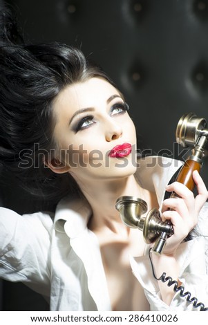 Portrait of passionate young brunette woman with bright makeup in white blouse holding retro gold telephone receiver looking away standing on black background, vertical photo