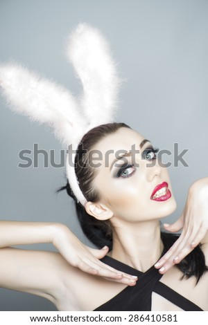 Playful sexy young woman with bright makeup and fluffy pink bunny ears standing on light grey backgound, vertical picture