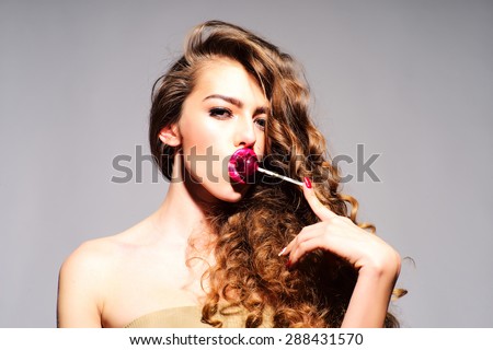 Revishing playful young undressed woman with curly hair and bright pink lips holding with finger purple round lollipop in mouth looking forward standing on grey background, horizontal picture