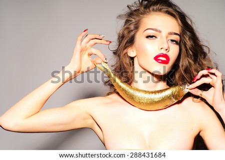 Inviting young naked woman with curly hair holding golden fish as necklace looking forward standing on grey background, horizontal picture