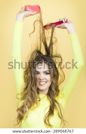 Portrait of smiling cute young lady with long curly hair in yellow blouse and blue jeans holding two pink hair brushes  standing on yellow background, vertical picture