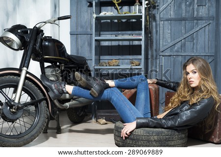 Tempting sexy young lady in black lace blouse and blue jeans lying near old fashioned motorcycle in garage interior on grey wooden wall background, horizontal picture