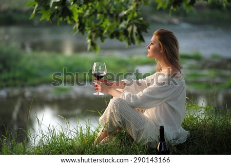 Alluring young lady in white clothes sitting on river bank in calm drinking red wine on natural sunset background, horizontal picture