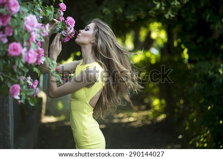 Beautiful young blonde lady with bright makeup and curly hair in yellow dress smelling lush bush of pink rose, horizontal picture