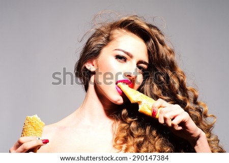 Heavenly young girl with curly hair and bright pink lips holding and biting bread roll looking forward standing on grey background, horizontal picture