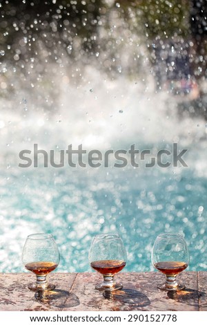 Amber alcoholic beverage in three glass bocals stnading in line on stone rim of fountain on water splashes background copyspace, vertical picture