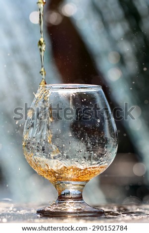 Amber alcoholic drink poured in bocal stnading on stone rim of fountain on water splashes background, vertical picture