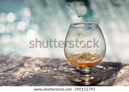 Amber alcoholic drink poured in glass stnading on stone rim of fountain on water splashes background copyspace, horizontal picture