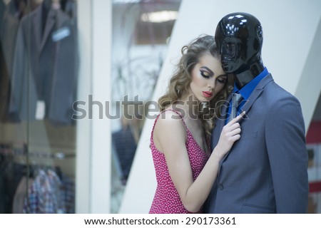 Misterious young girl with bright makeup and curly hair standing with male mannequin in formal clothes on shopping background, horizontal picture