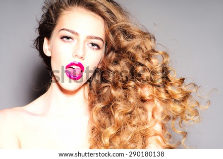 Amazing sexy young girl with curly hair licking her bright pink lips with tongue looking forward standing on grey background, horizontal picture