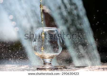 Amber alcoholic liquor poured in glass stnading on stone rim of fountain on water splashes background, horizontal picture