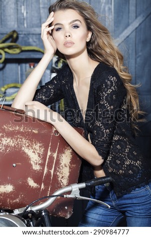 Pensive sexy young woman in black lace blouse standing near old fashioned aged brown suitcase in garage on grey wooden wall background, vertical picture