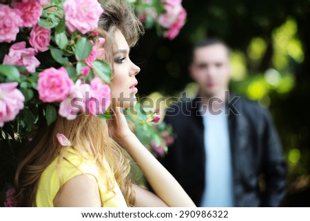 Passionate young blonde woman with bright makeup and curly hair in yellow dress smelling lush bush of pink rose and man in background, horizontal picture