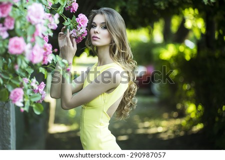 Pretty young blonde woman with bright makeup and curly hair in yellow dress touching lush bush of pink rose, horizontal picture