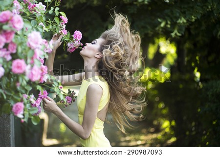 Attractive young blonde lady with bright makeup and curly hair in yellow dress smelling lush bush of pink rose, horizontal picture
