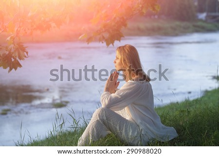 Misterious young woman in white clothes sitting on river bank in calm on natural sunset background, horizontal picture